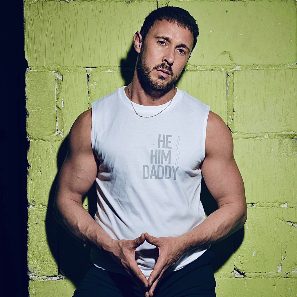 A person wearing a white sleeveless vest with the text 'HE HIM DADDY' on it, standing against a green textured background.