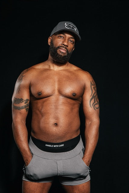 handsome black man wearing gay underwear under grey shorts reading Handle with Care on the waistband 